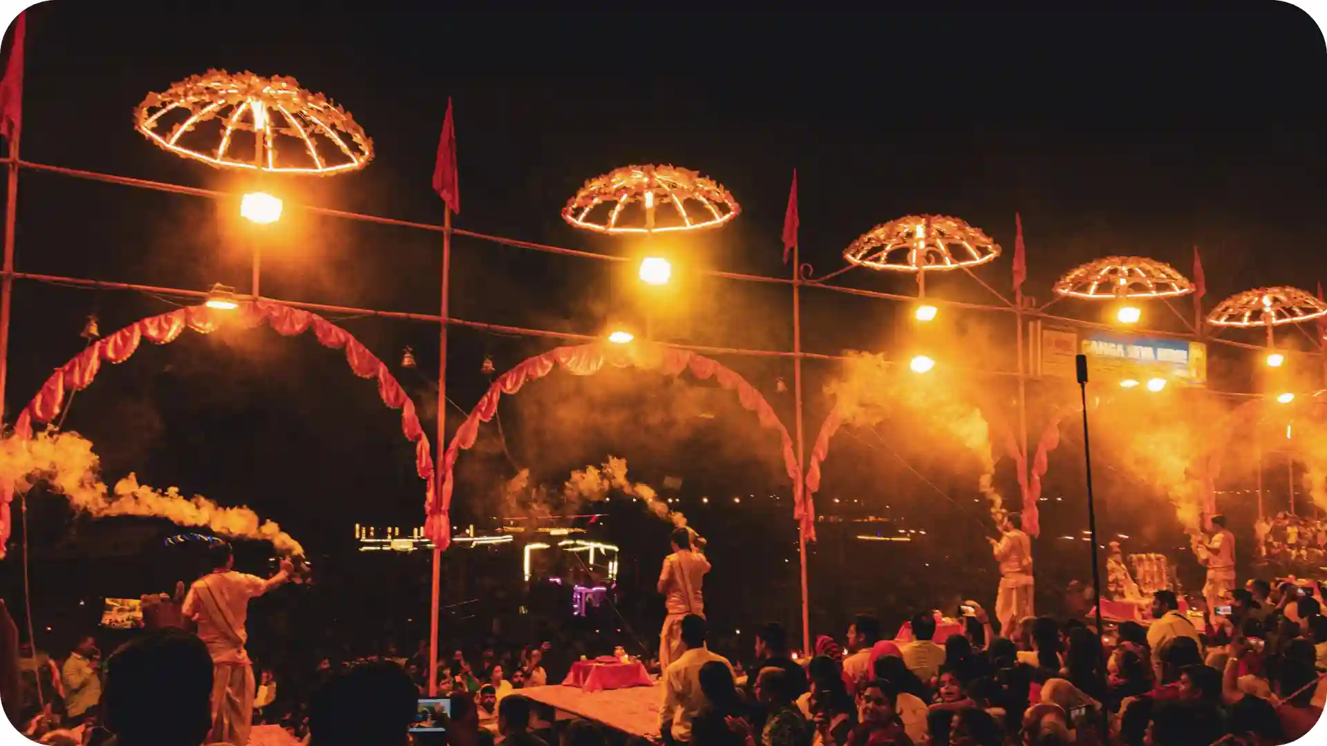 Devotees gathered on Varanasi ghats watching priests perform Ganga Aarti night ceremony with flames, incense and illuminated arches along the sacred river