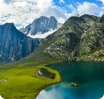 Turquoise alpine lake surrounded by lush green meadows and dramatic snowy Himalayan peaks under blue sky, showcasing Kashmir trekking and high-altitude scenic landscapes