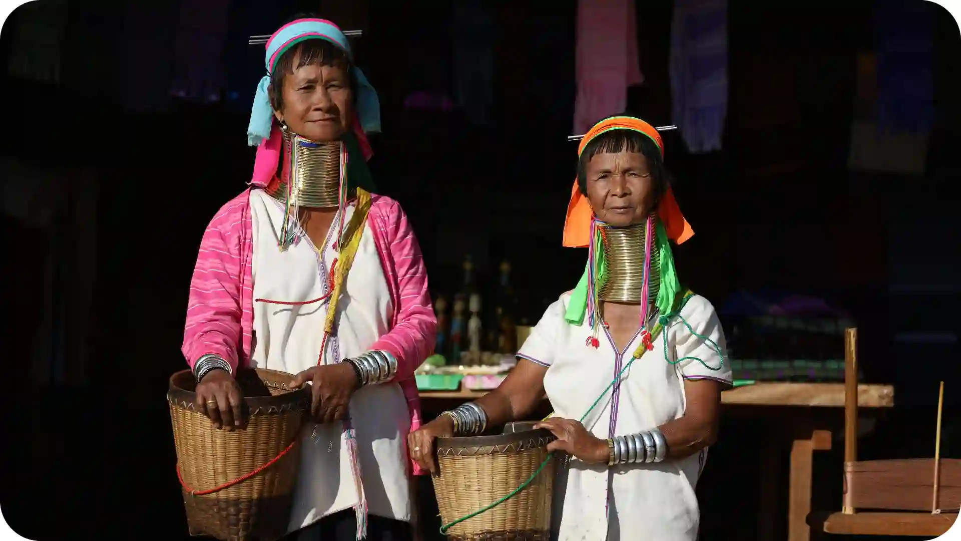Two tribal women with neck rings and woven baskets standing in traditional dress, featured on Ghumora’s Northeast India tribal trails adventure tour page